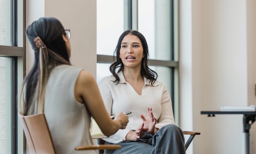 Two professional women sit across from each other in front of tall windows, one speaking with expressive hand gestures while the other listens attentively.
